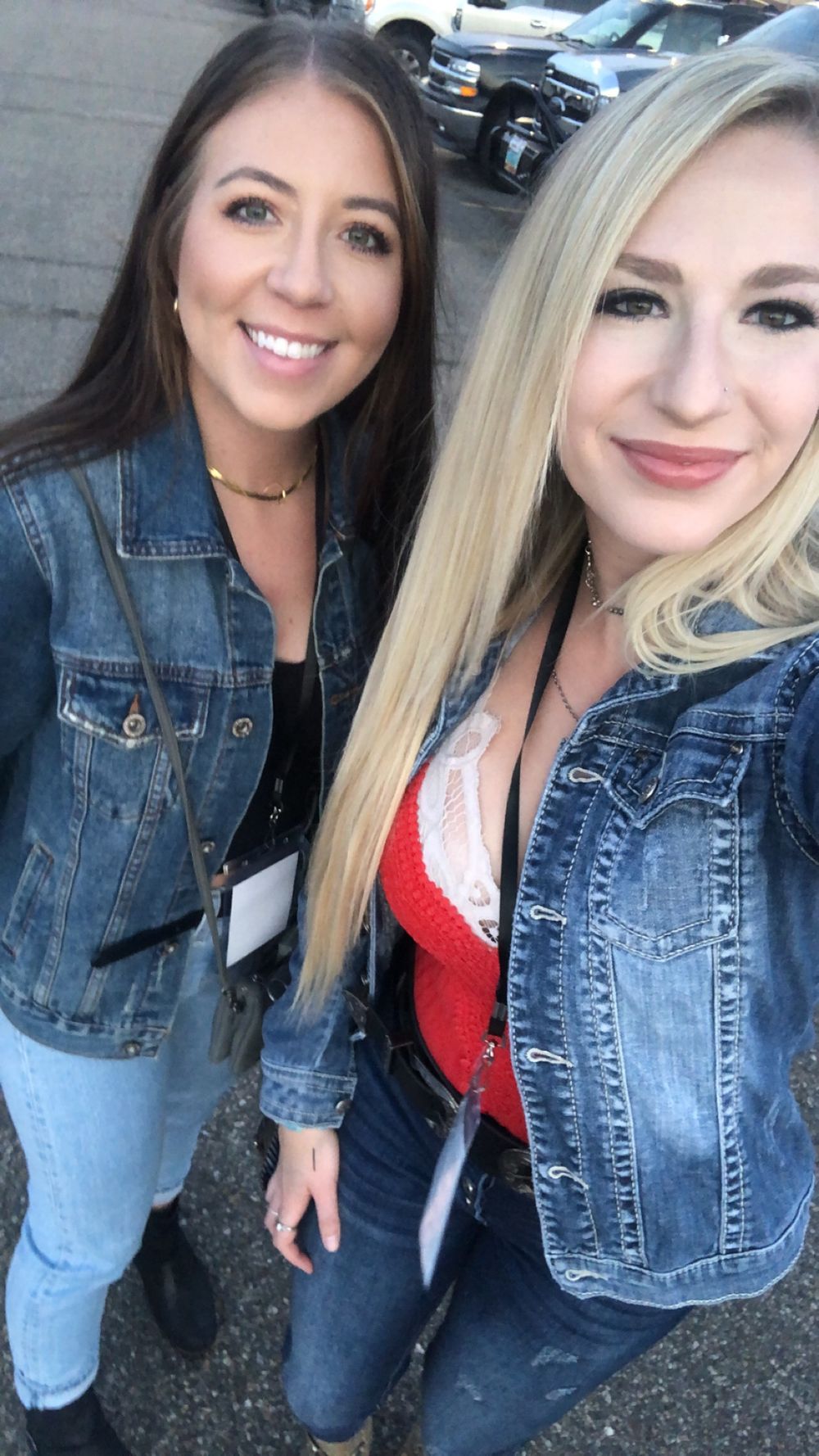 Two smiling women in matching denim jackets pose for a selfie outdoors near a parking area.
