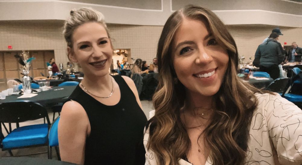 Two women smiling at a formal event, one in a black dress and one in a floral top, with banquet tables in the background.