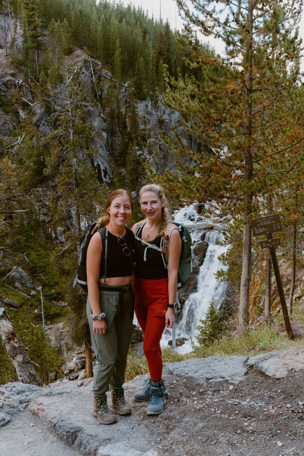 Two women hikers with backpacks smile on a mountain trail with a waterfall and pine trees in the background.