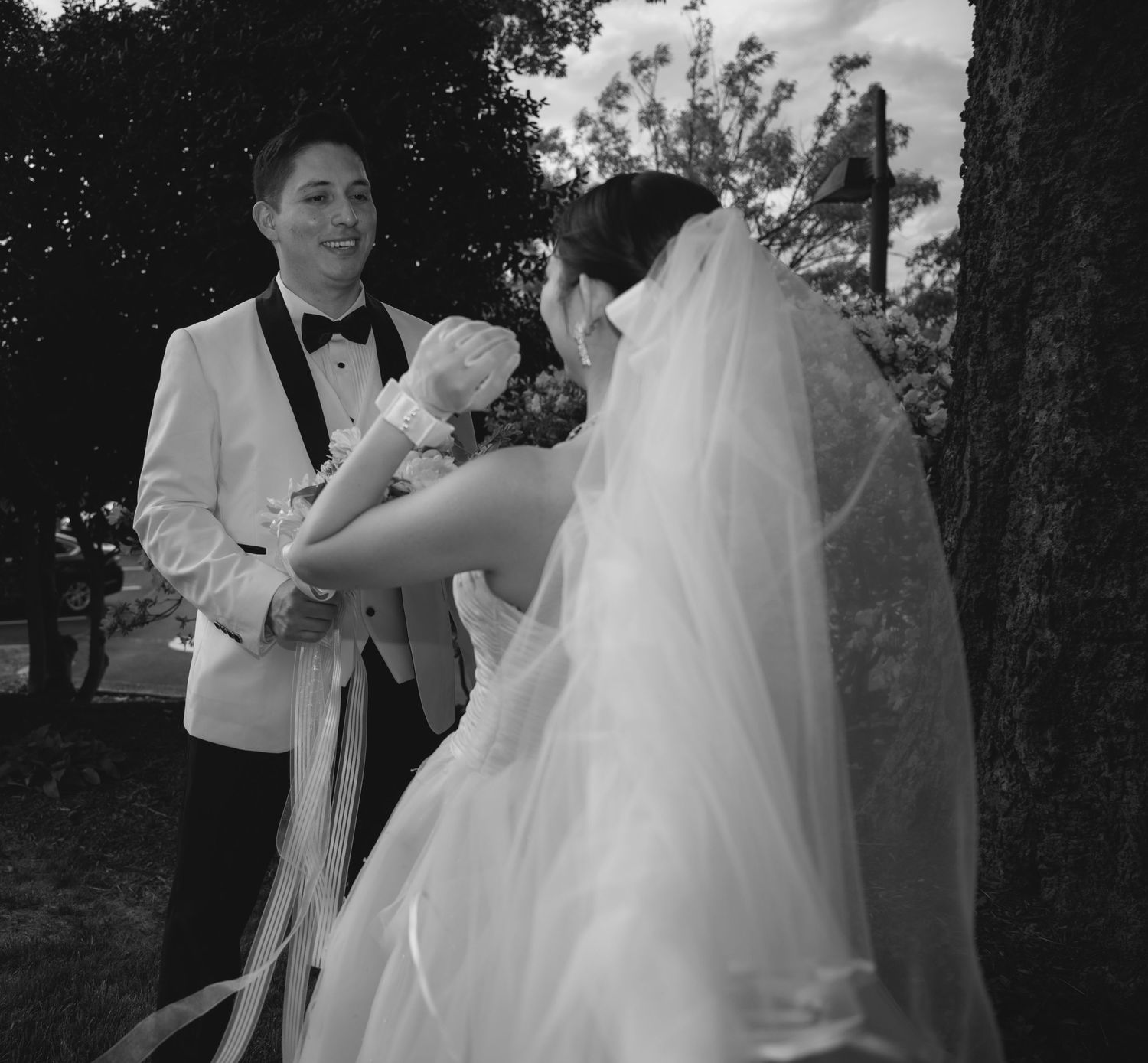 Black and white photo of a smiling groom in white tuxedo with bride in veil and wedding dress near a tree.