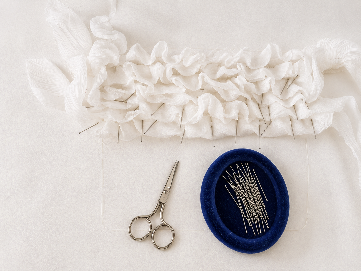 White ruffled fabric pinned to surface with straight pins, small scissors, and a blue pin cushion with sewing pins.
