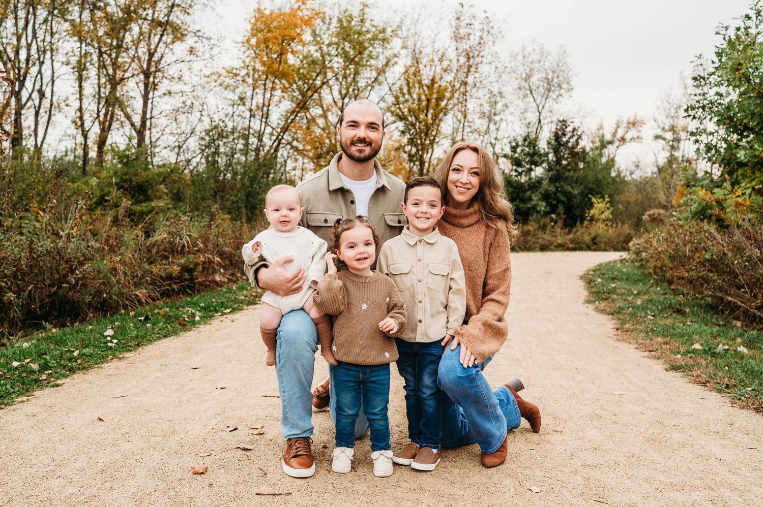 A smiling family of four poses on a gravel path surrounded by autumn trees during a fall outdoor photo session.