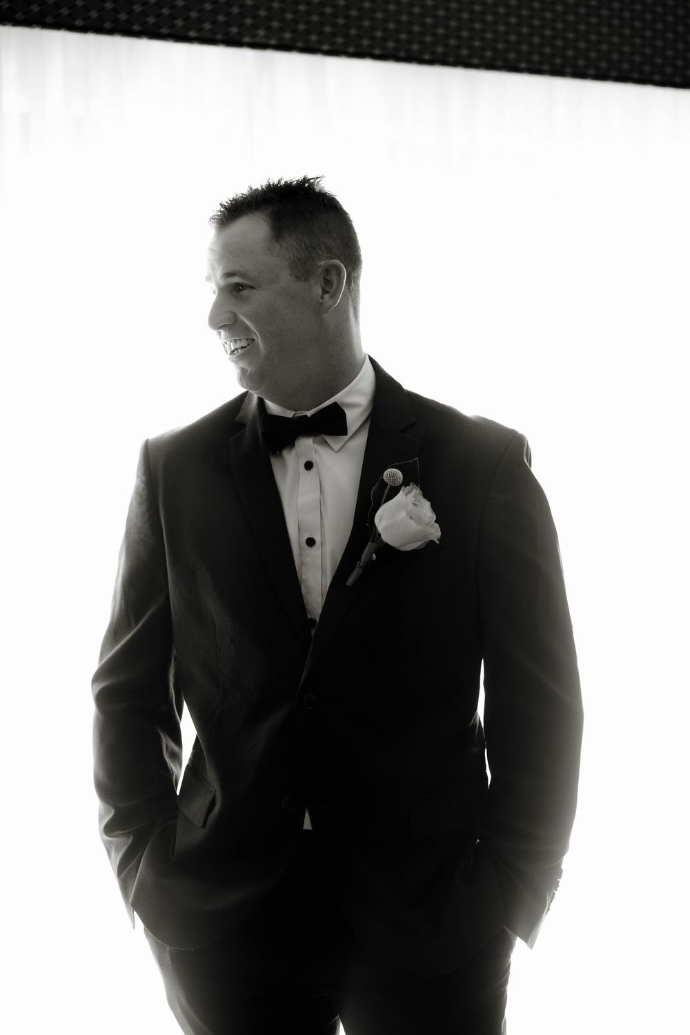 Groom in black tuxedo with boutonniere poses confidently in black and white wedding photo.