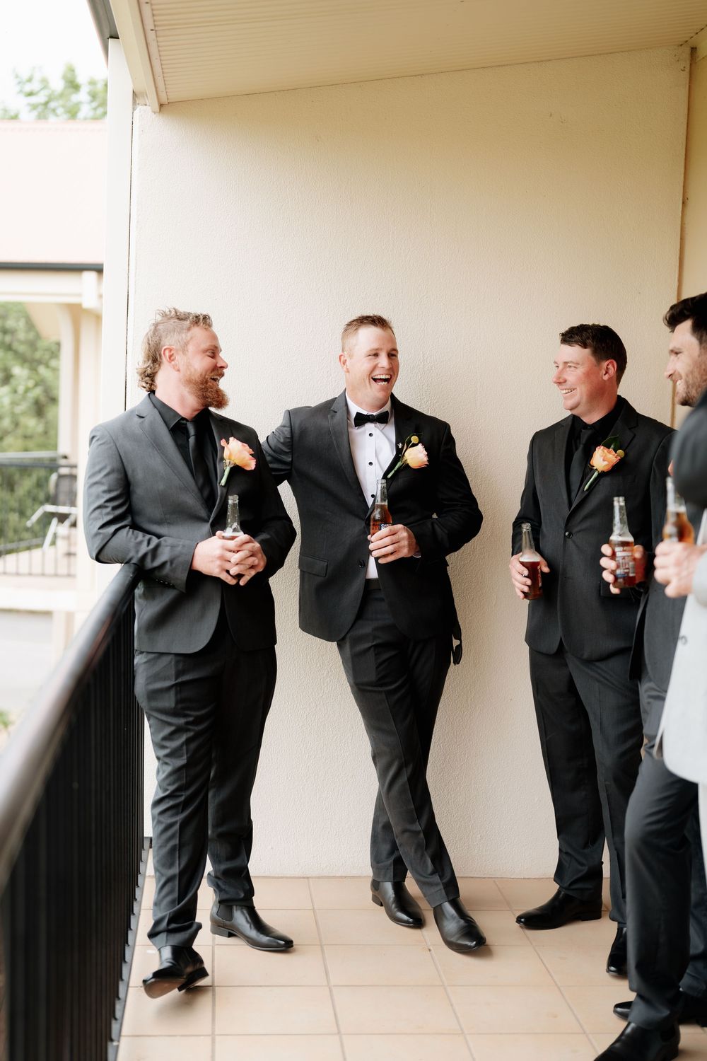 Groom and groomsmen in dark suits laugh together on balcony holding drinks before wedding.