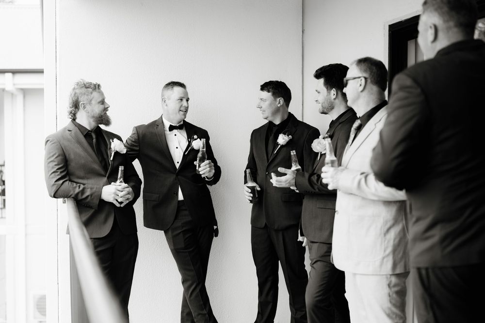 Groomsmen in black tuxedos socializing and holding drinks before a wedding ceremony in black and white.