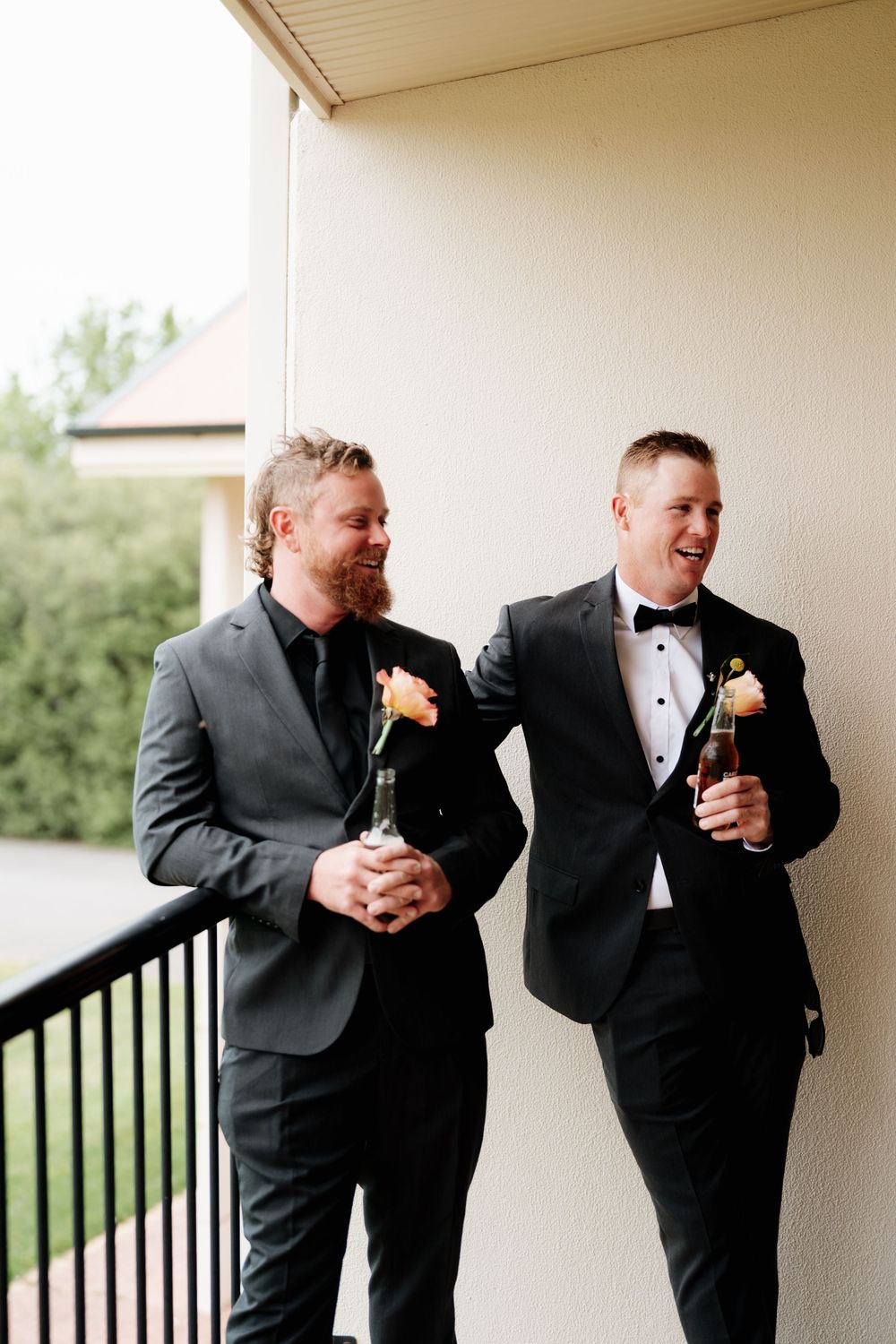 Two groomsmen in black tuxedos with floral boutonnieres laugh on a balcony holding beer bottles at a wedding.