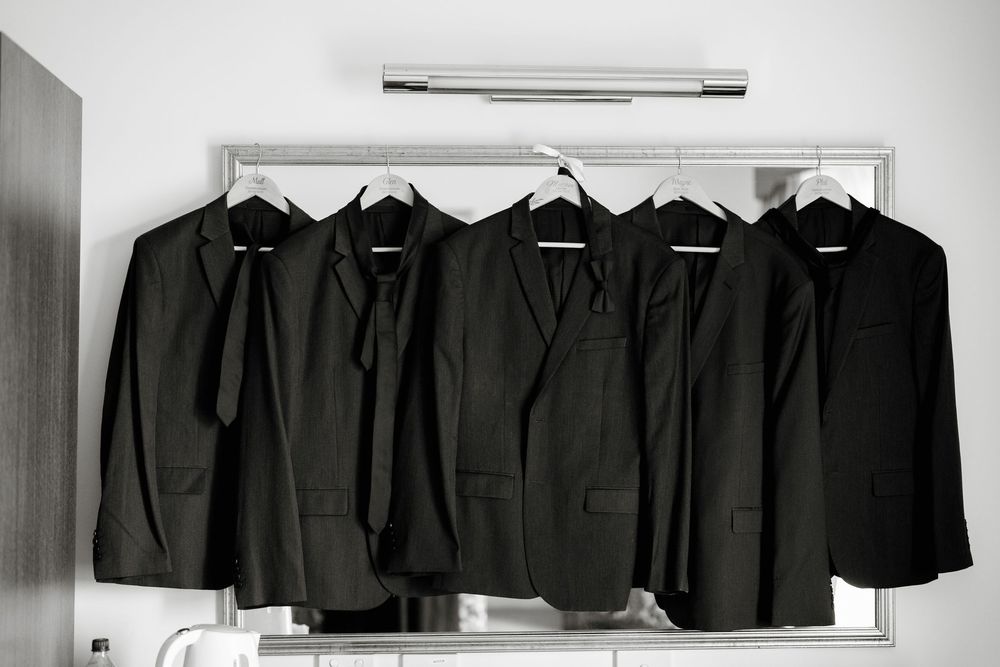 Five black suits on hangers displayed on a rack against a white wall.