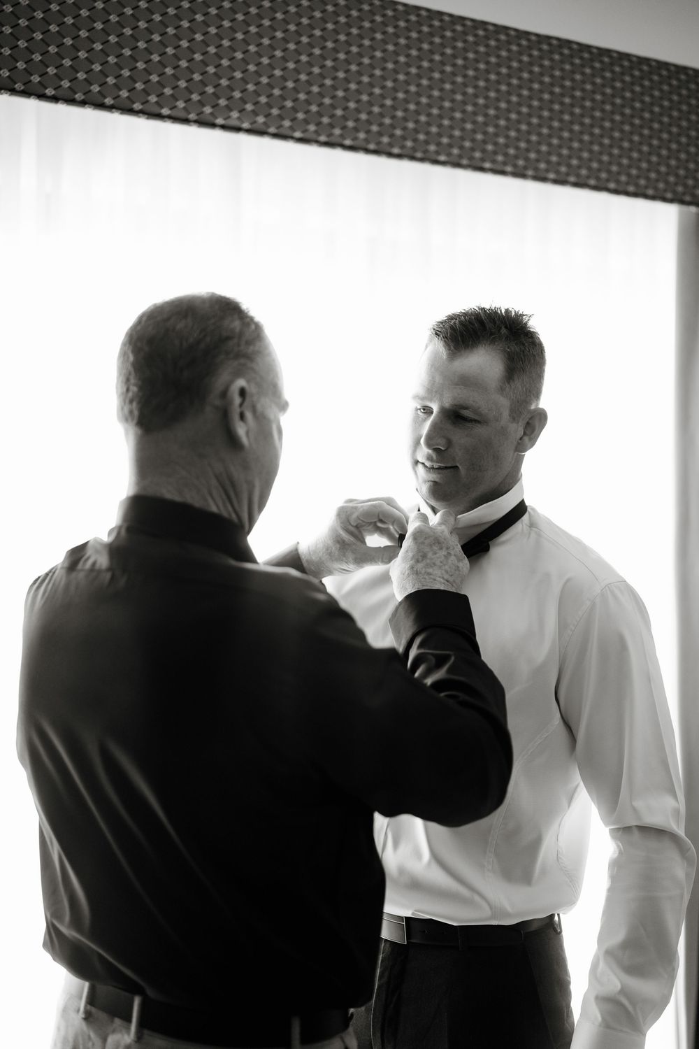 Black and white photo of older man adjusting bow tie of younger groom before wedding ceremony.
