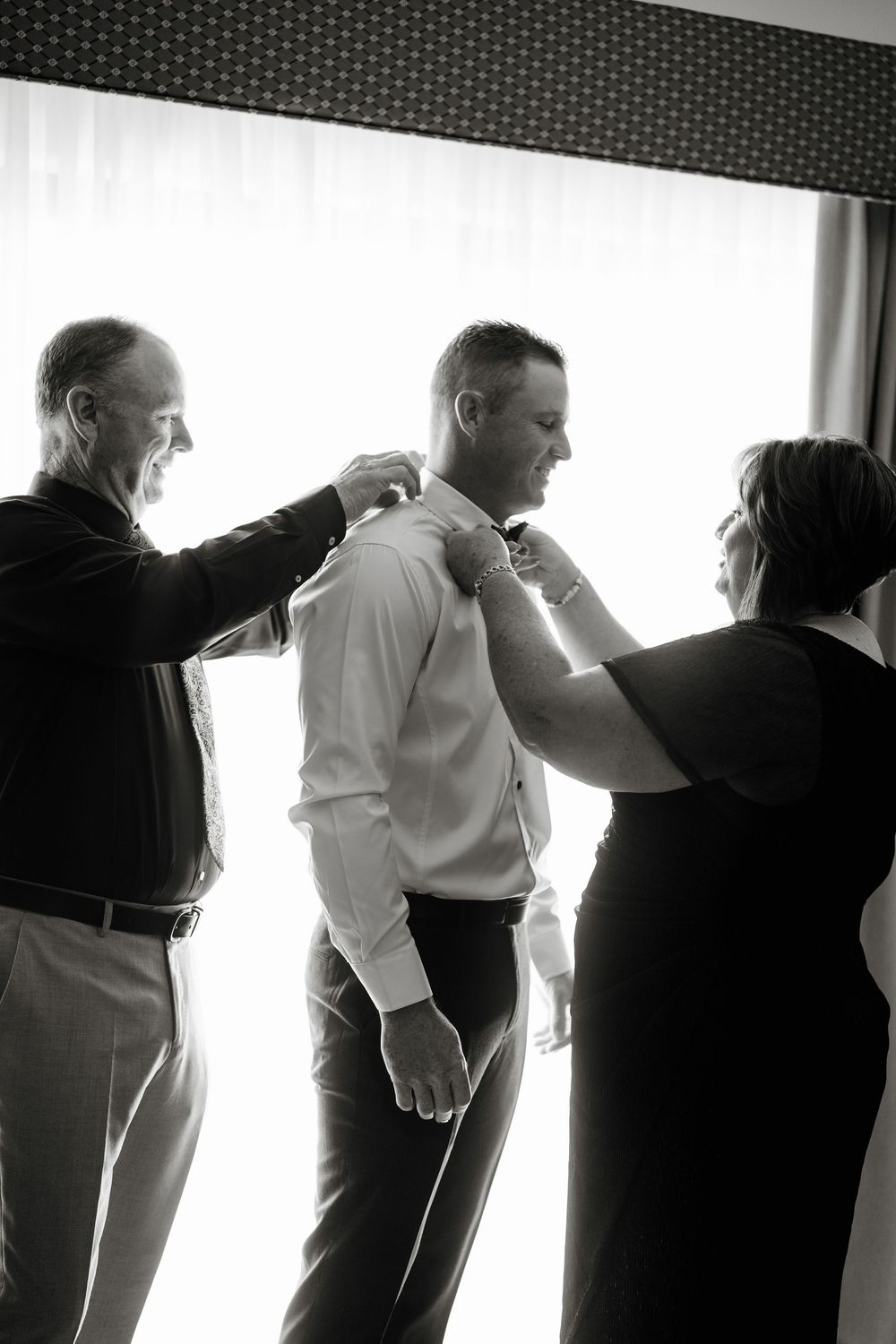 Black and white photo of parents helping groom get dressed before his wedding ceremony.