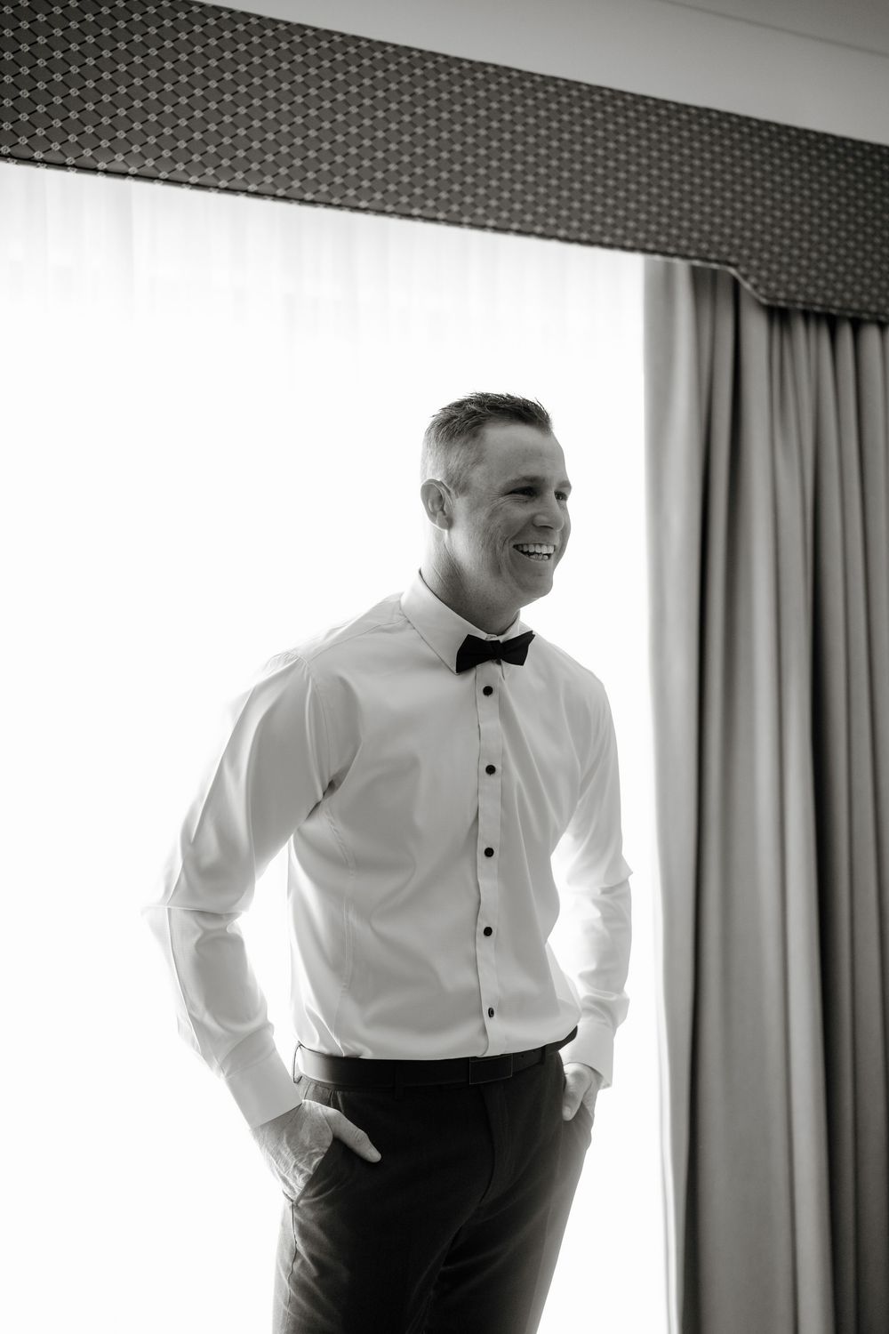 Smiling groom in white dress shirt and black bow tie standing by a bright window in black and white photo.