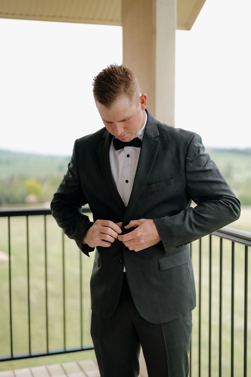 Groom in dark tuxedo with bow tie buttoning jacket on outdoor balcony with green landscape background.