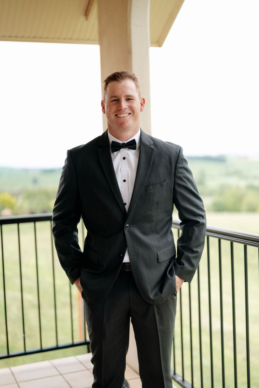 Smiling groom in dark tuxedo with bow tie stands on balcony with green landscape backdrop.