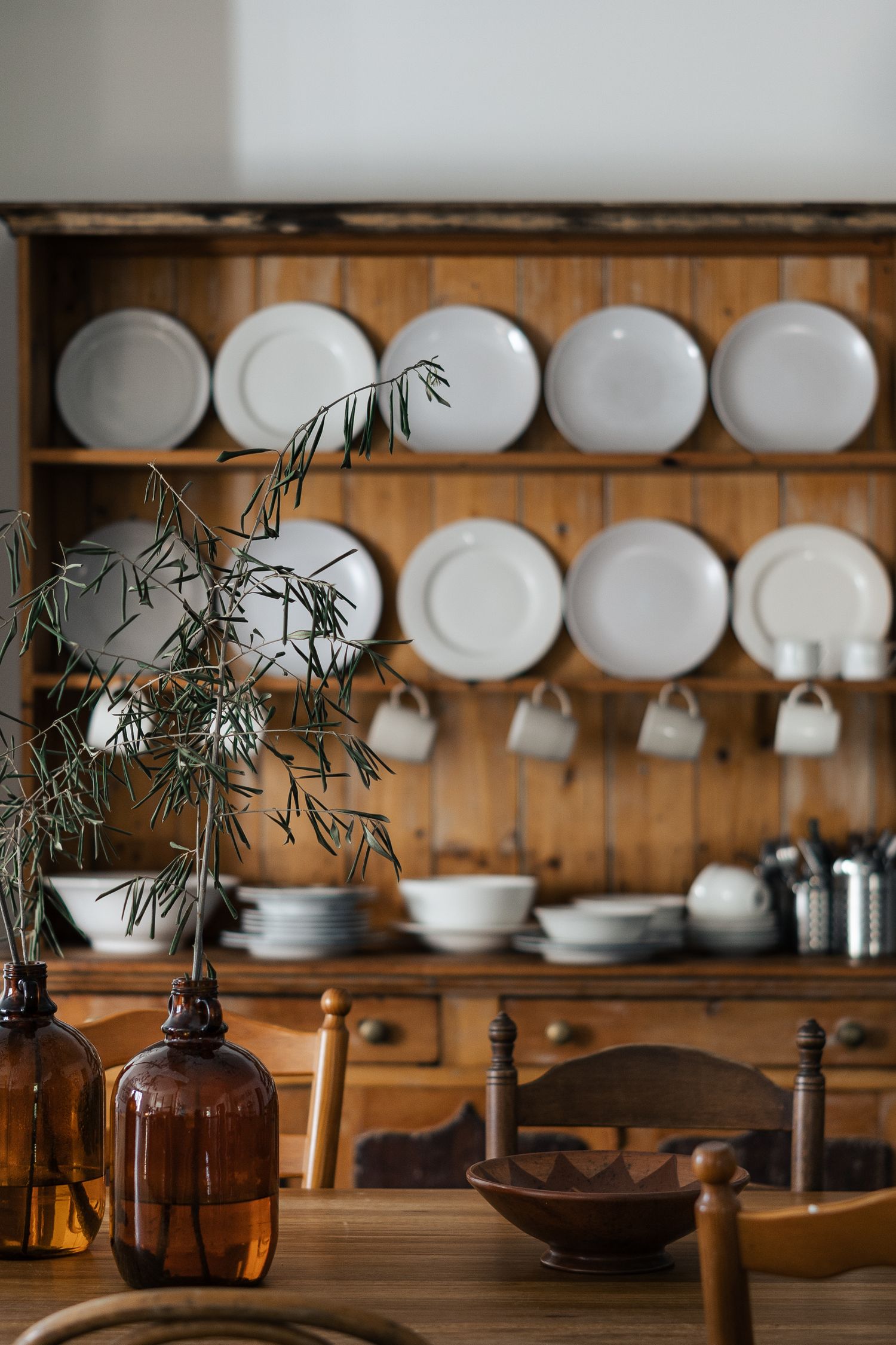 Rustic wooden hutch displaying white plates and cups on shelves with amber bottles and plant in foreground.

Piccadilly House