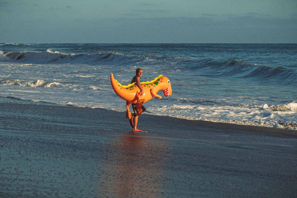 Person carrying a large orange inflatable float on a sandy beach with ocean waves in the background.