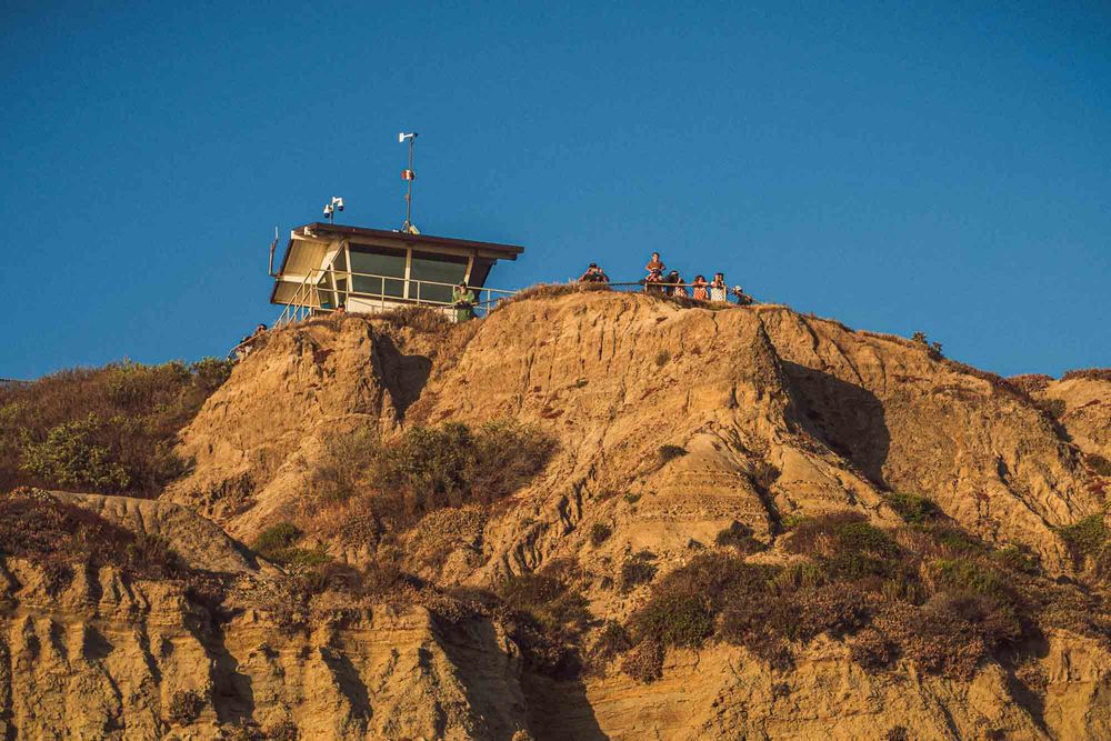 A rustic lifeguard station perched atop rugged golden cliffs against a clear blue sky.