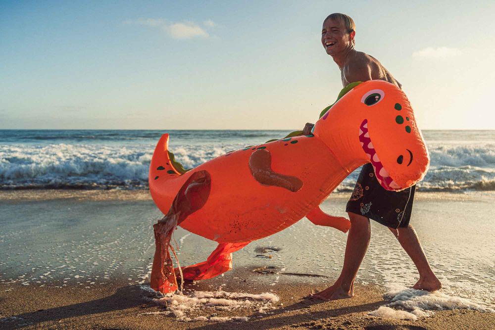 Boy carrying an orange inflatable dinosaur toy on a sunny beach with ocean waves in the background.