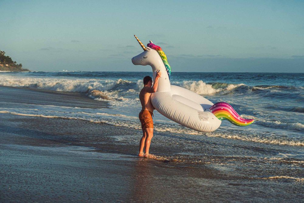 Person carrying a large inflatable unicorn float on a sandy beach with ocean waves in the background.