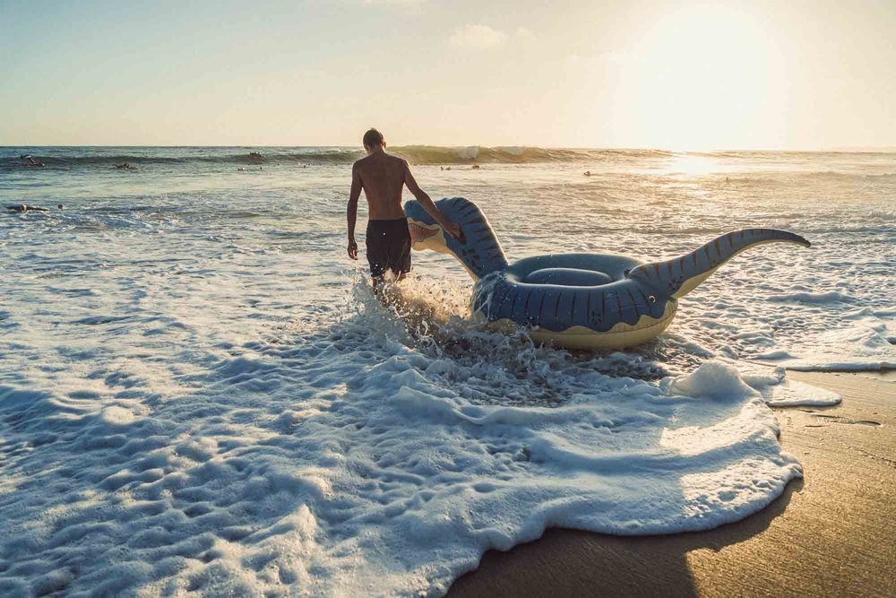 Man dragging an inflatable dinosaur float into ocean waves at sunset on a sandy beach.