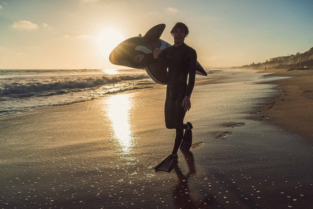 Surfer in wetsuit and fins carrying a bodyboard along a sandy beach at golden sunset.