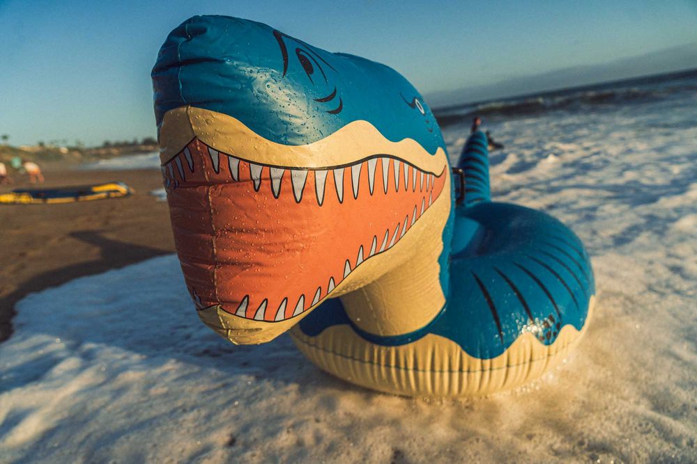 Close-up of a colorful inflatable shark pool float on a sandy beach with ocean waves.