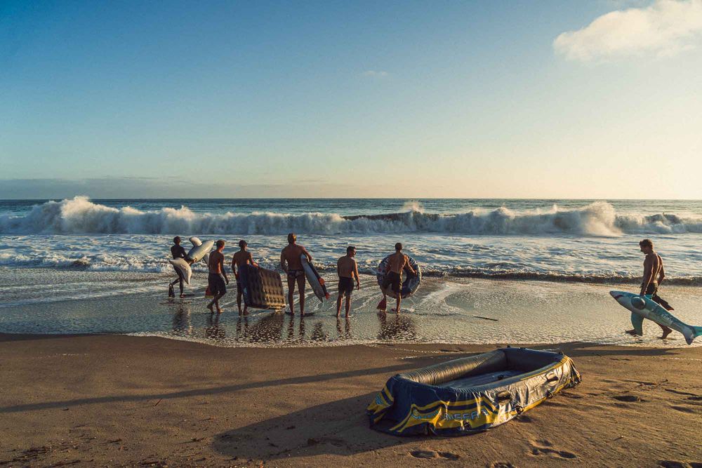 Group of surfers walking into ocean waves at sunset with an inflatable kayak on sandy beach.