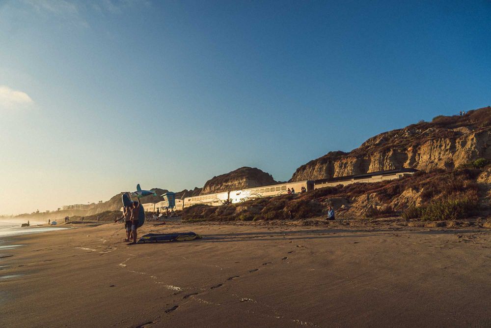 People walking on a wide sandy beach at sunset with rocky cliffs in the background.