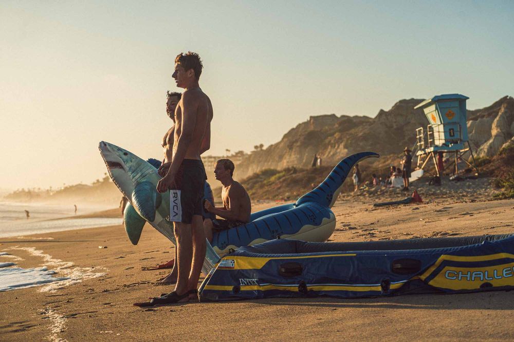 Two surfers stand on a sunny beach at golden hour holding colorful inflatable shark floaties near sandy cliffs.