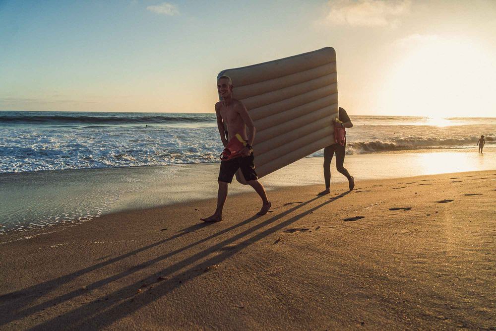 Two people carrying an inflatable air mattress along a sunny beach at golden sunset.