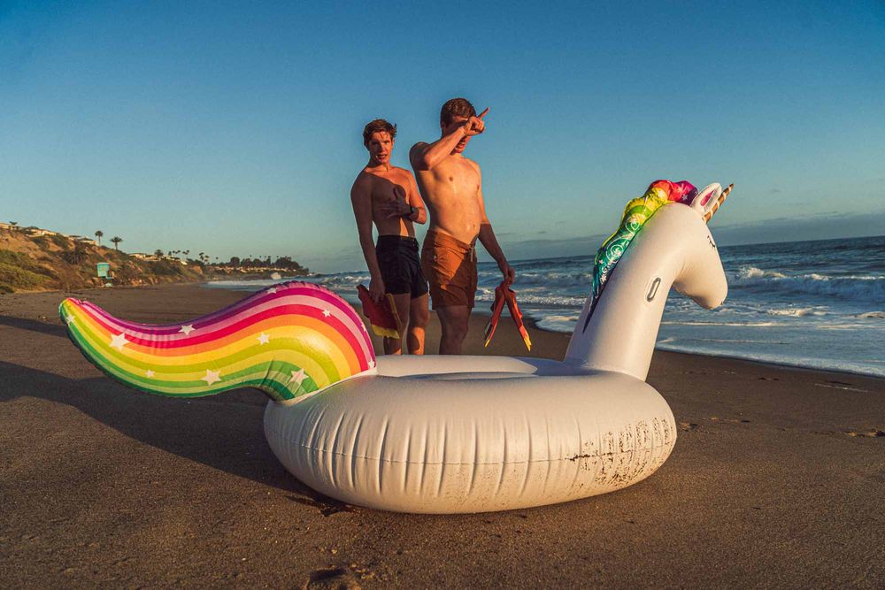 Two men stand on a sunny beach beside a large inflatable unicorn pool float with a rainbow tail.