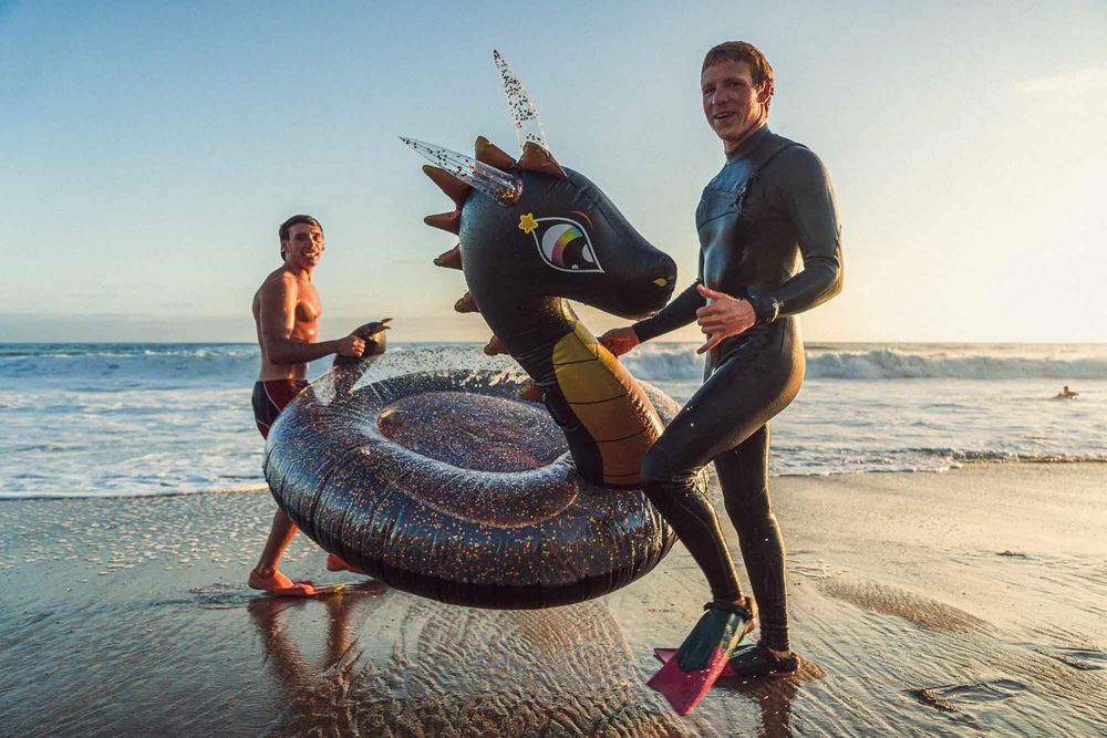 Two men on a sunny beach holding a dragon-shaped inflatable ring near ocean waves.