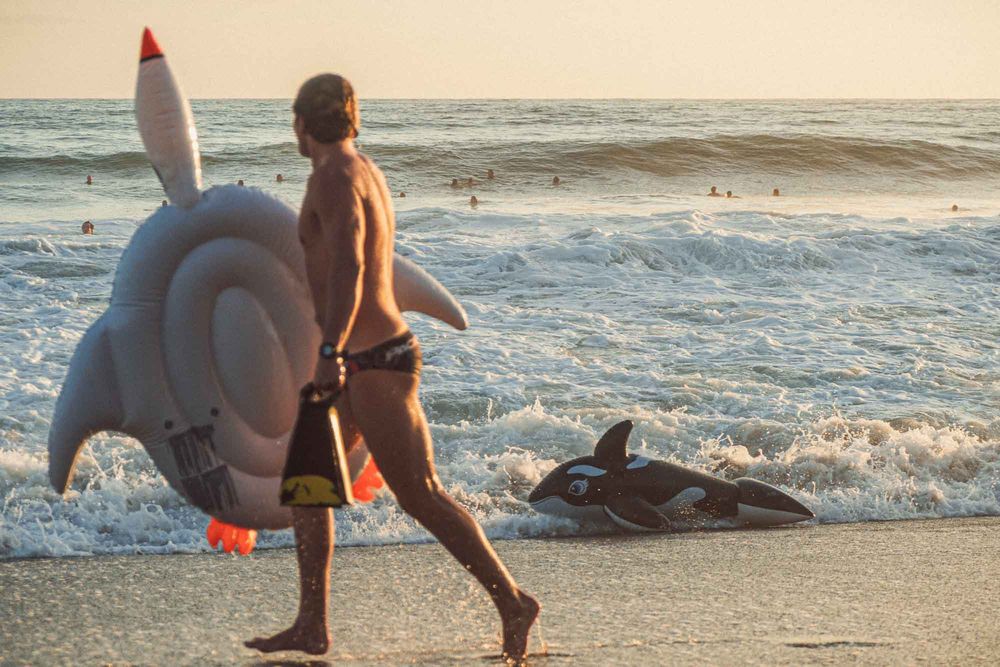 Man walking on beach carrying inflatable shark float at sunset with ocean waves behind him.