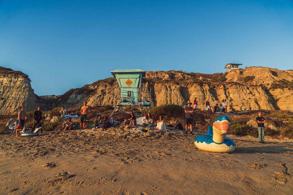 Beach crowd gathers near a colorful lifeguard tower at golden hour with inflatable pool toys on sandy shore.
