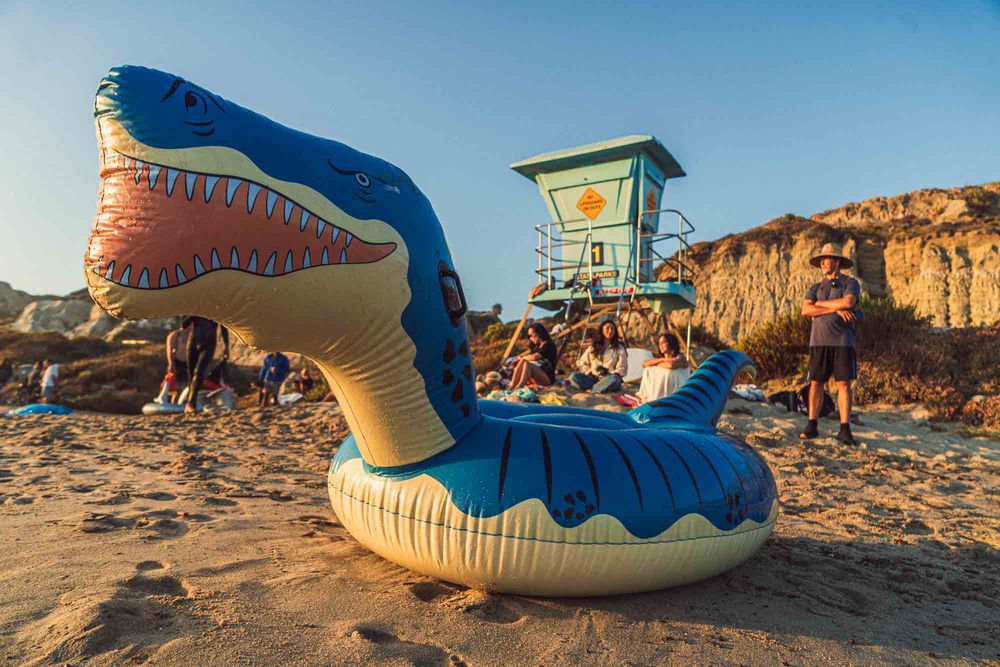 Large inflatable T-Rex dinosaur float on a sandy beach with people relaxing near a lifeguard tower at sunset.