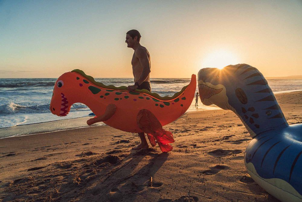 Man on a beach at sunset holding inflatable dinosaur floats with ocean waves and golden light in the background.