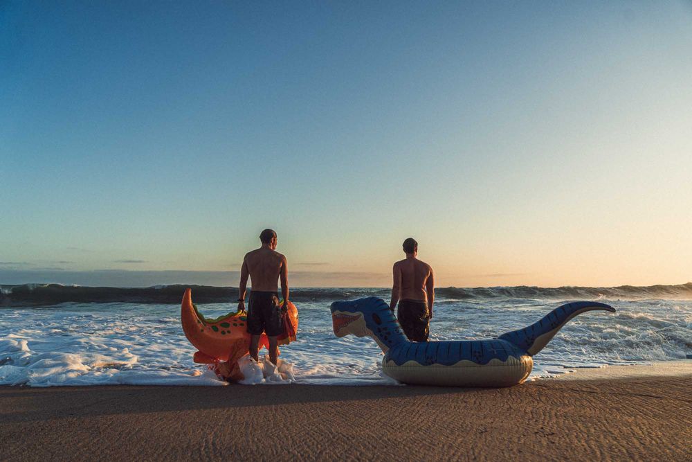 Two shirtless men stand at ocean's edge at sunset holding inflatable dragon and shark pool floaties in surf.