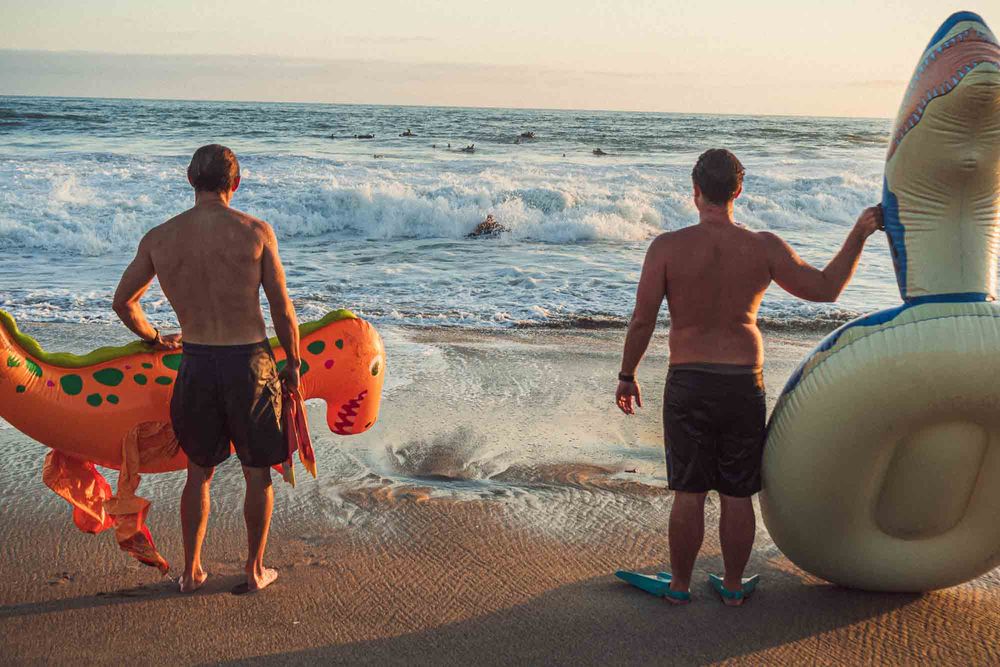 Two shirtless men standing on beach at sunset holding colorful inflatable animal pool floats.