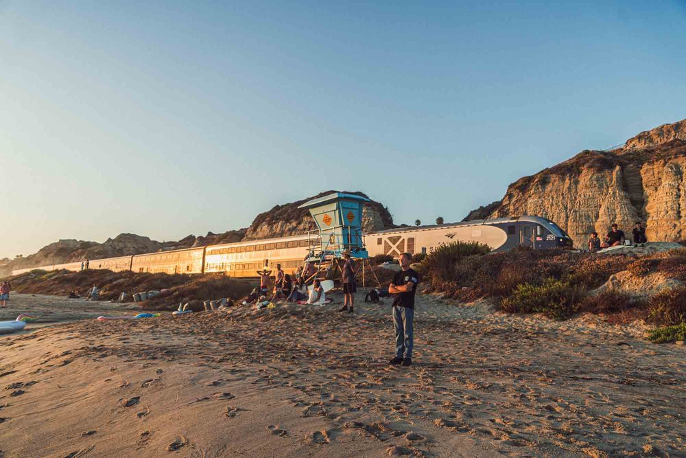 Person standing on sandy beach near a lifeguard tower and cliffs at golden hour sunset.