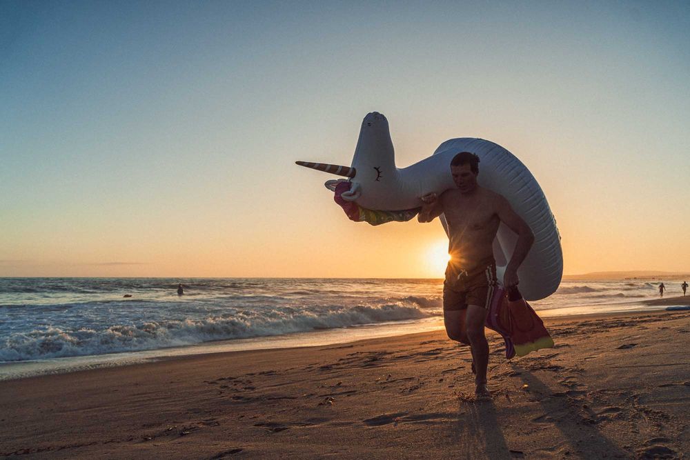 Man running on beach at golden sunset carrying a large inflatable airplane float.