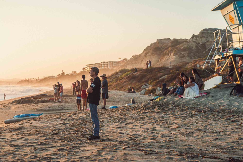 People gather on a sandy beach at golden hour with a lifeguard tower and coastal cliffs in the background.