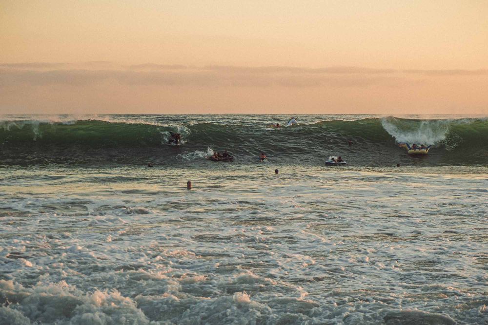 Surfers riding large ocean waves at sunset with golden sky and white foam crashing on the shore.
