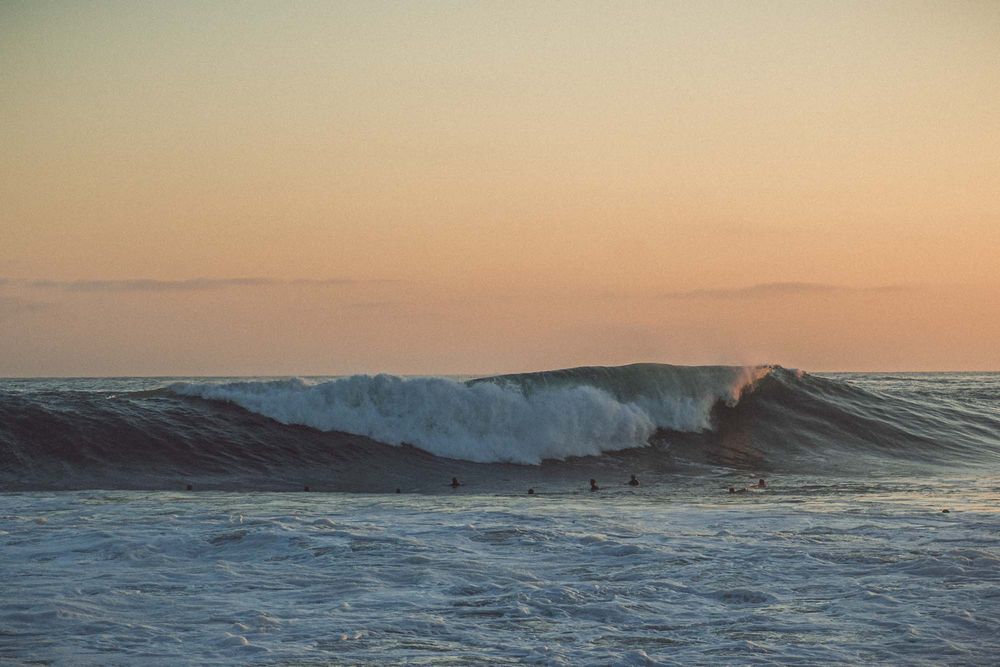 Surfers wait in lineup beyond powerful breaking ocean wave at golden sunset on a dramatic coastline.