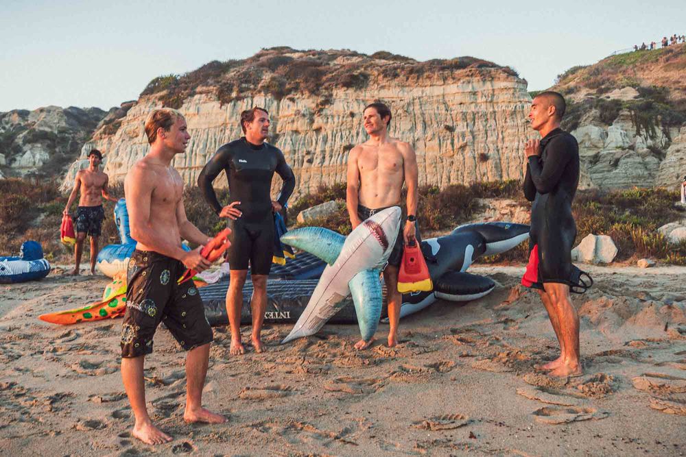 Group of surfers in wetsuits chatting on a sandy beach with colorful surfboards and rocky cliffs behind.