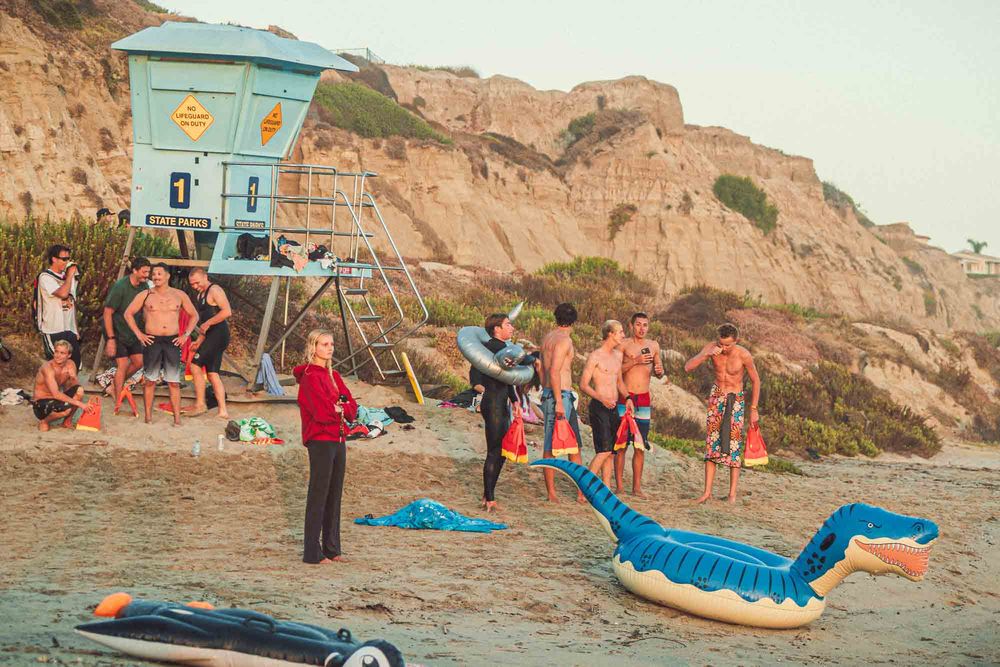 Beach scene with surfers near a blue lifeguard tower, inflatable toys on sand, rocky cliffs in background.