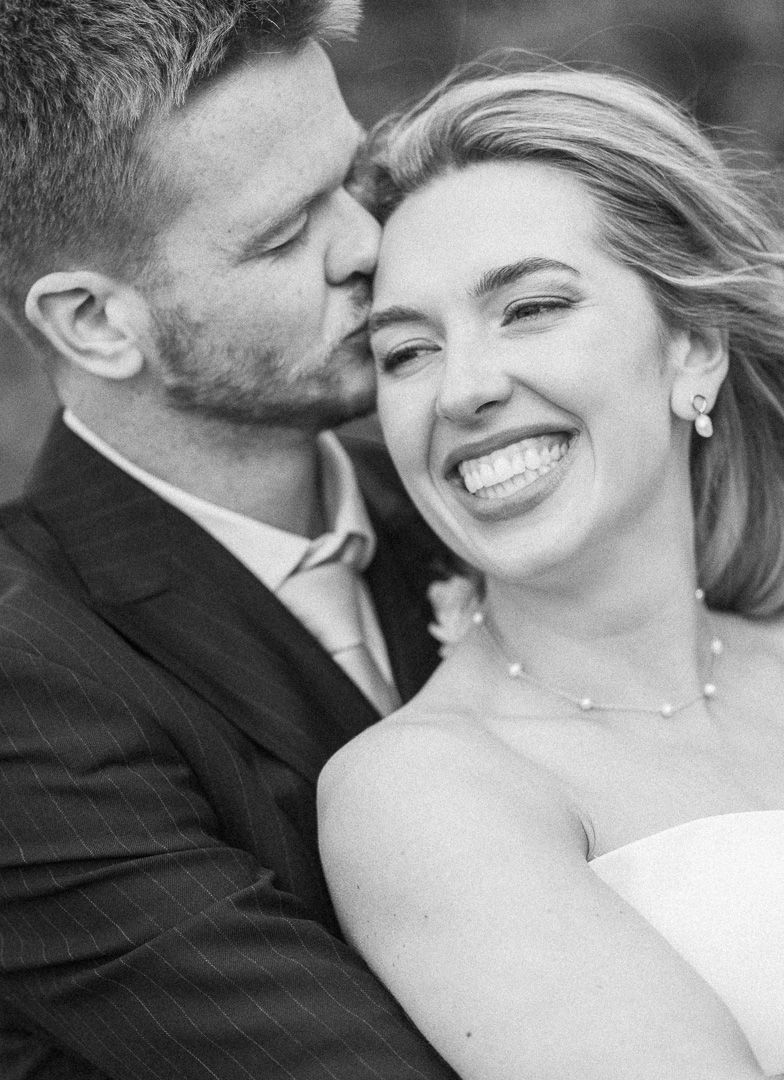Black and white wedding photo of smiling bride and groom sharing a tender kiss on their special day.