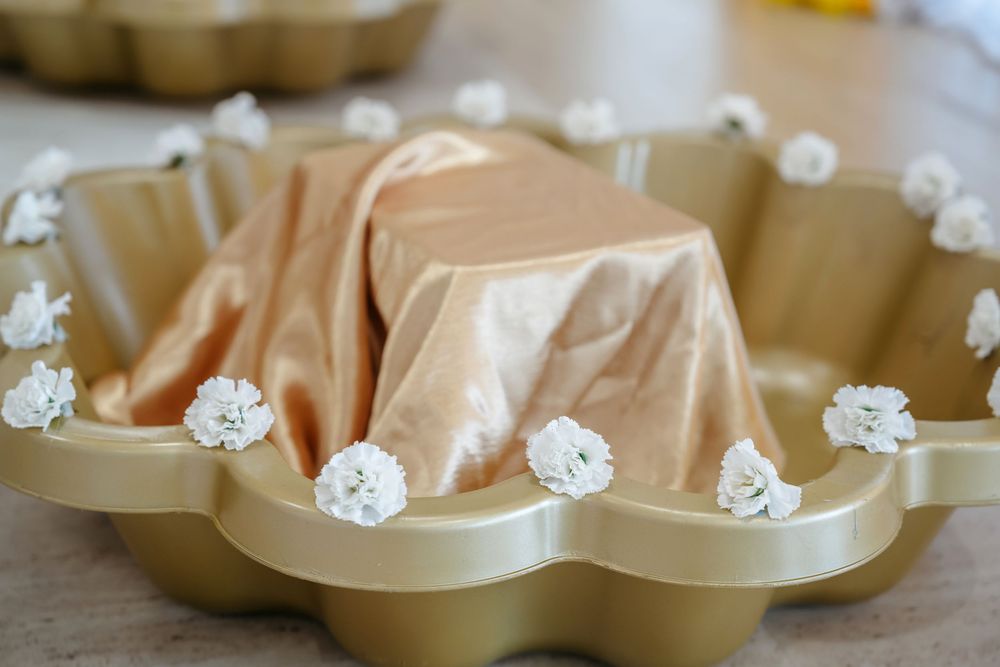 Gold bundt cake pan decorated with white flower accents and a golden satin cloth draped over a cake.