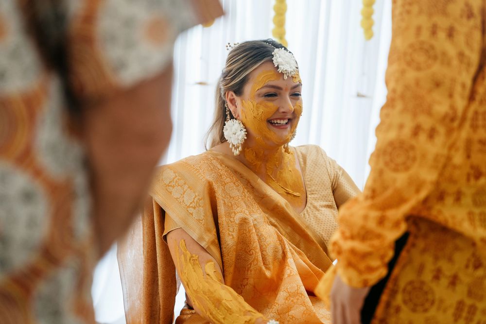 Smiling bride in golden sari with turmeric haldi ceremony makeup and floral hair accessories.