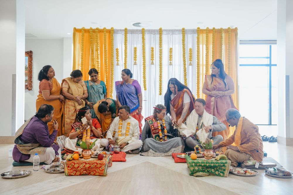 A large Indian family gathers for a traditional Hindu wedding ceremony with colorful attire and ritual offerings indoors.