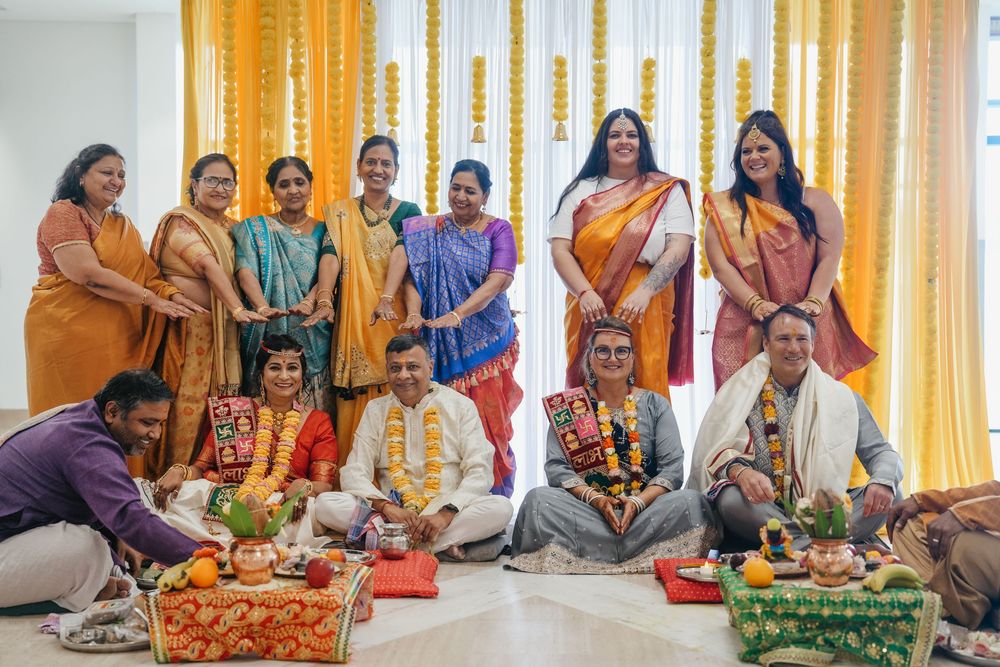 Traditional Indian wedding ceremony with family group photo, colorful sarees, floral decorations, and ritual offerings on a decorated stage.