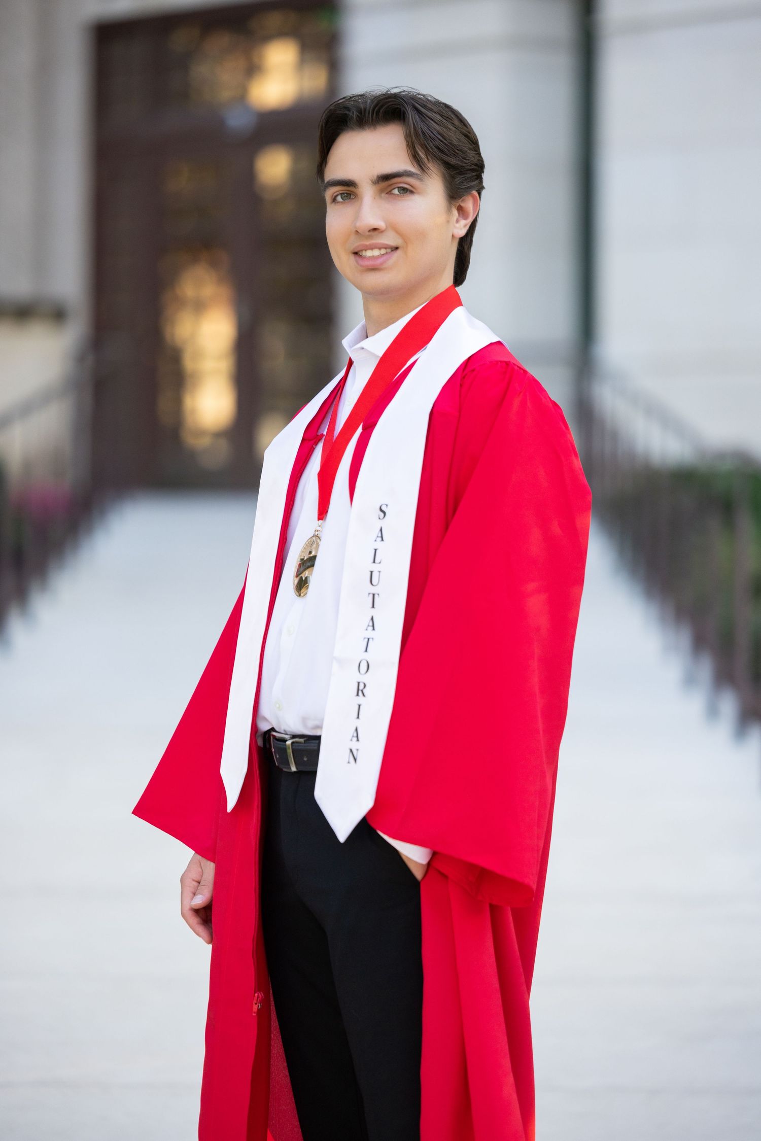 Young man in red graduation gown and cap smiling outdoors, holding mortarboard, wearing honor cords at graduation ceremony.