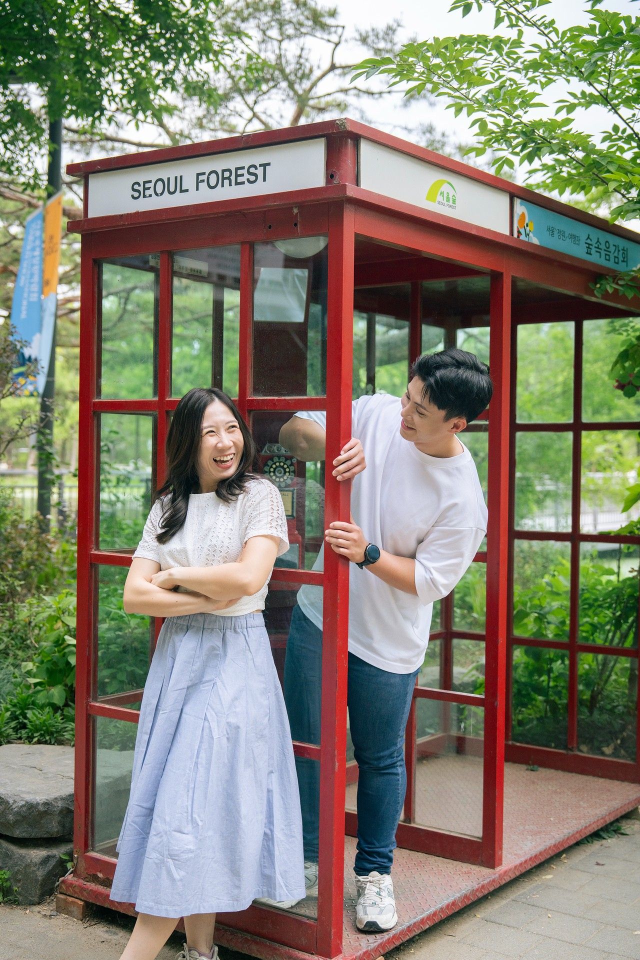 A laughing woman in a blue dress and a man in white pose at a red Seoul Forest phone booth in South Korea.